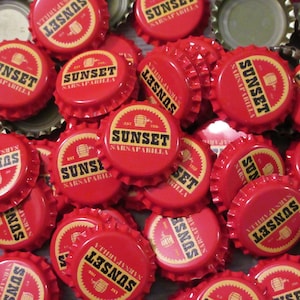 May include: A collection of red metal bottle caps featuring a circular design with the text SUNSET SARSAPARILLA in black lettering, a small barrel icon, and the text EST 1956. The caps have a serrated edge and a silver-colored interior lining.