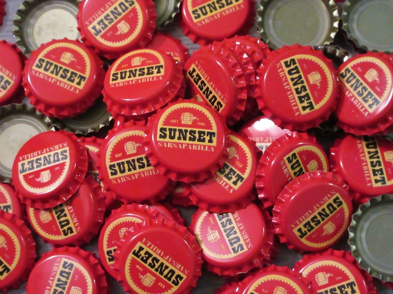 May include: A collection of red metal bottle caps featuring a circular design with the text SUNSET SARSAPARILLA in black lettering, a small barrel icon, and the text EST 1956. The caps have a serrated edge and a silver-colored interior lining.