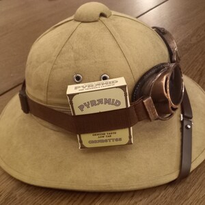 May include: A khaki-colored pith helmet with a brown band and attached goggles. A pack of "PYRAMID" cigarettes is secured to the hat. The goggles have brown frames and lenses. The hat has two small eyelets on the front.