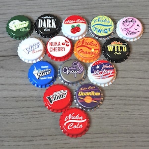 May include: A collection of fourteen metal bottle caps featuring various colorful graphic designs and logos, including Nuka-Cola, Nuka-Cherry, Nuka-Orange, Nuka-Wild, Nuka-Grape, Nuka-Victory, Nuka-Quantum, Nuka-Twist, Nuka-Dark, Vim, and Vim Quartz.