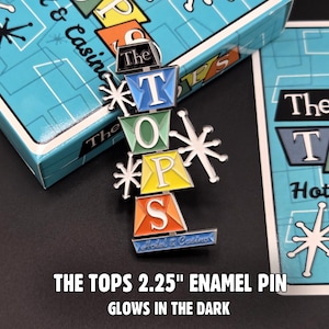 May include: An enamel pin with the words "The TOPS" in a stacked design with colourful blocks and starburst accents. The pin is 5.7 cm and glows in the dark. The pin is next to a box and a card with a similar design.