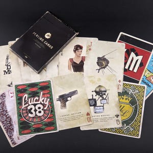 May include: A deck of playing cards with a black box labelled "PLAYING CARDS". The cards display vintage illustrations, including a woman, a gun, and the words "Lucky 38". The cards are spread out on a black surface.