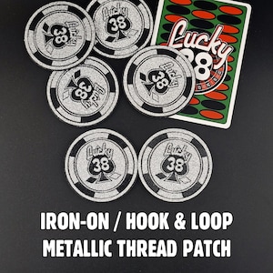May include: Six circular patches featuring a silver metallic thread design of a spade with the number 38 and the word Lucky. One rectangular patch displays a green and red pattern with the text Lucky 38. The text IRON-ON / HOOK & LOOP METALLIC THREAD PATCH is visible.