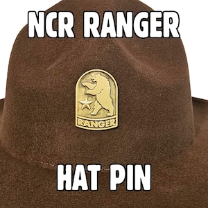 Può includere: Un cappello marrone con la scritta "NCR RANGER HAT PIN" in lettere bianche. Una spilla color oro con un orso e una stella è attaccata al cappello. Il cappello ha un'ampia tesa e una corona arrotondata.