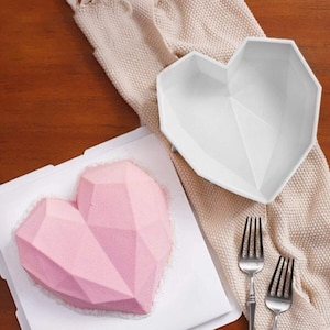 May include: A pink geometric heart-shaped cake on a white square plate. A white heart-shaped mold is on a beige fabric next to the cake. Two silver forks are on the table.