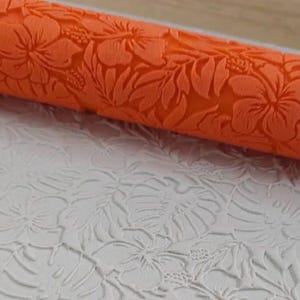 Puede incluir: Rodillo de amasar naranja con un diseño floral. El diseño presenta flores de hibisco y hojas. El rodillo se utiliza para crear un patrón en relieve en fondant o masa.