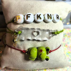 May include: Three handmade bracelets. The top bracelet has white beads with gold letters spelling "PKNR". The middle bracelet has a clear charm with a heart cutout. The bottom bracelet features a green frog charm. All bracelets are on a cream-colored pillow.