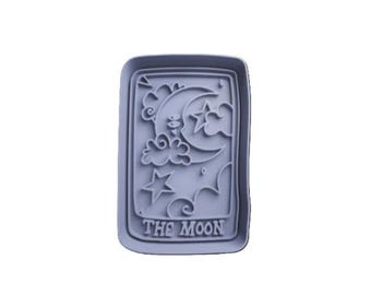 May include: A light gray, rectangular cookie cutter featuring a crescent moon, stars, and clouds. The words "THE MOON" are at the bottom. The design is a representation of the tarot card "The Moon".