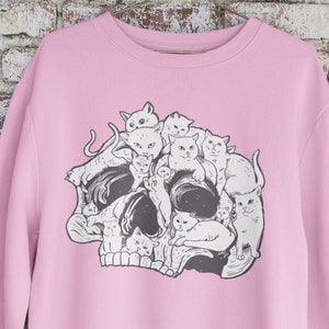 May include: A pink sweatshirt with a graphic of a skull made up of cats. The cats are all different sizes and are arranged in a way that makes them look like they are forming the skull.