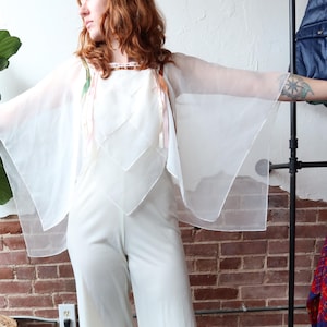 Könnte beinhalten: Weißer Jumpsuit mit einem transparenten, kapuzenartigen Überzug. Der Jumpsuit hat weite Beine und einen quadratischen Ausschnitt. Der Überzug ist an den Schultern befestigt und fällt über die Arme. Die Person trägt beige spitz zulaufende Schuhe. Ziegelwand im Hintergrund.