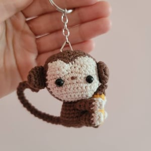 May include: A brown crocheted monkey keychain holding a yellow banana. The monkey has black eyes and a white face.