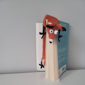 May include: A crocheted fox bookmark in orange with black eyes and a white nose. The bookmark is hanging from a book titled "The Childhood Adventures of Winnie the Pooh and The House at Pooh Corner."