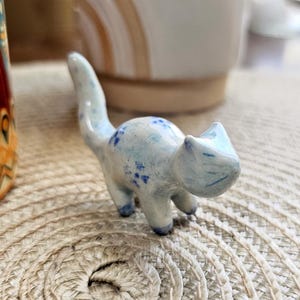 May include: A small, ceramic cat figurine in a light blue color with darker blue spots. The cat has a curved tail and is posed in a walking position. The figurine is on a woven surface.