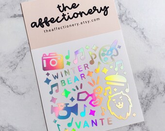 May include: A sheet of iridescent holographic stickers featuring a variety of designs, including musical notes, stars, a paint palette, a camera, a trumpet, a bear, and the words "Winter Bear" and "Vante".