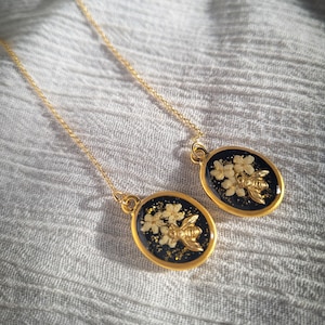 Puede incluir: Pendientes colgantes dorados. Cada pendiente presenta un colgante ovalado con fondo negro, flores secas y una abeja dorada. Los pendientes cuelgan de finas cadenas doradas, ofreciendo un diseño elegante e inspirado en la naturaleza.