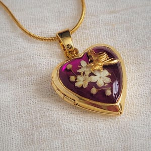 May include: A gold-coloured heart-shaped locket necklace with a purple interior. The locket contains dried white flowers and a gold-coloured bee. The necklace chain is also gold-coloured.