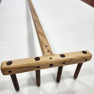 May include: A wooden rake with a long handle and a T-shaped grip. The rake head has four tines and is decorated with dark circular inlays. The wood has a natural grain and a light brown color.