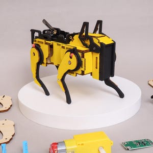 May include: A yellow and black wooden robot dog model stands on a white platform. The robot dog has articulated legs and a black head. Various components, including a motor, circuit board, and wooden pieces, are scattered around the platform, suggesting a DIY kit.