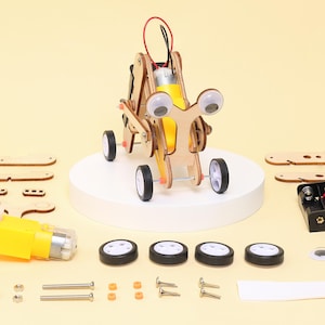 May include: A wooden robot toy with googly eyes, a yellow motor, and four black and white wheels. The robot is assembled on a white platform, with additional unassembled parts and hardware visible.