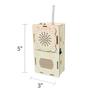 May include: A wooden toy radio, 5 inches tall and 3 inches wide, with a speaker, antenna, and switches. The radio has a natural wood finish with a speaker and a series of holes. The antenna is a silver coil. The radio has a green and red button.