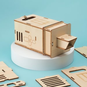 May include: A wooden box with a slot on the top and a small opening on the side. The box is made of light wood and has a honeycomb pattern on the front. The box is sitting on a white pedestal.