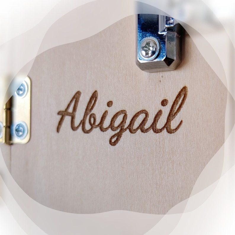 K&ouml;nnte beinhalten: Nahaufnahme einer Holzoberfl&auml;che mit dem Namen "Abigail" in kursiver Schrift graviert. Das Holz ist hell, die Gravur ist dunkelbraun. Metallscharniere und ein Verschluss sind sichtbar.