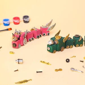 May include: A whimsical steampunk-style train set in pink and green, adorned with metallic gears, wings, and decorative elements. Paint pots and a paintbrush are visible, suggesting a DIY craft project. The train cars are connected, and various small hardware pieces are scattered around.