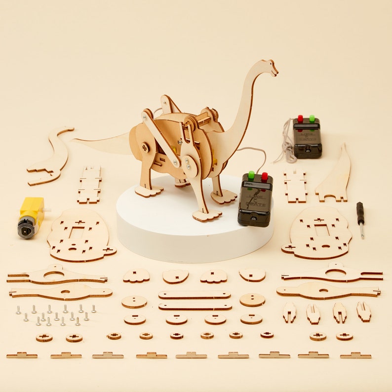 Puede incluir: Un kit de juguete de dinosaurio de madera con control remoto. El kit incluye una variedad de piezas, incluyendo el cuerpo del dinosaurio, las patas, la cola y la cabeza. El dinosaurio est&aacute; ensamblado sobre una base blanca. El kit tambi&eacute;n incluye un destornillador y una peque&ntilde;a herramienta amarilla.