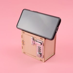 K&ouml;nnte beinhalten: Eine hellbraune Holzkiste mit Zahlenschloss, die ein schwarzes Smartphone h&auml;lt. Die Box hat silberne Beschl&auml;ge. Das Telefon ist schwarz und auf der Oberseite der Box angewinkelt. Der Hintergrund ist rosa.