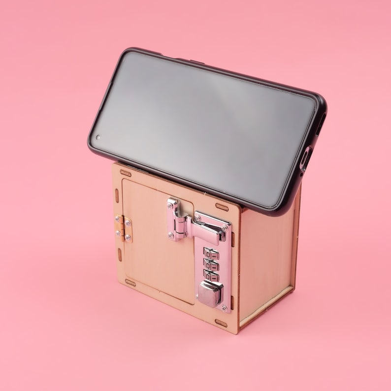 K&ouml;nnte beinhalten: Eine hellbraune Holzkiste mit Zahlenschloss, die ein schwarzes Smartphone h&auml;lt. Die Box hat silberne Beschl&auml;ge. Das Telefon ist schwarz und auf der Oberseite der Box angewinkelt. Der Hintergrund ist rosa.