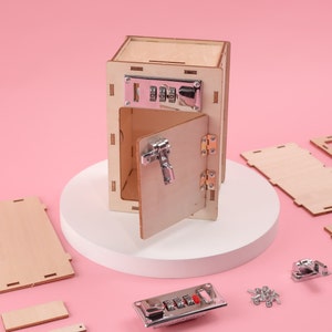 May include: A wooden toy safe with a combination lock and a hinged door. The safe is open and the door is ajar. The safe is on a white circular platform. There are several small wooden pieces and metal hardware pieces scattered around the safe.