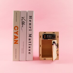 May include: A wooden box with a combination lock and a small door that is open. The box is sitting on a pink surface in front of three books. The books are titled "Henri Matisse", "Vulture", and "CYAN".