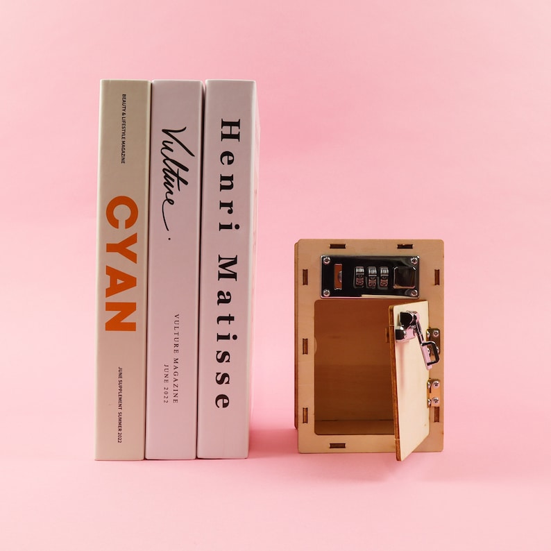 May include: A wooden box with a combination lock and a small door that is open. The box is sitting on a pink surface in front of three books. The books are titled "Henri Matisse", "Vulture", and "CYAN".