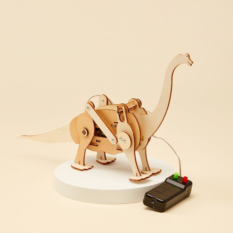 Puede incluir: Un dinosaurio de juguete de madera con un cuello largo y una cola que est&aacute; doblada hacia un lado. El dinosaurio est&aacute; sobre una plataforma blanca y tiene un mando a distancia con botones rojos y verdes. El dinosaurio est&aacute; hecho de madera y tiene un acabado natural.