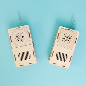 May include: Two wooden walkie-talkies with spring antennas, a speaker, and a microphone. The walkie-talkies are light brown and have a rectangular shape. The background is a solid light blue.