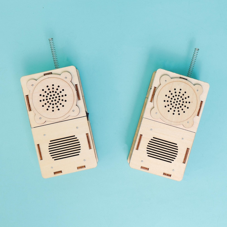 May include: Two wooden walkie-talkies with spring antennas, a speaker, and a microphone. The walkie-talkies are light brown and have a rectangular shape. The background is a solid light blue.