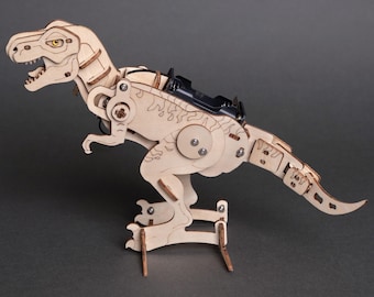 May include: A light-colored wooden model of a Tyrannosaurus Rex dinosaur featuring articulated joints, visible screws, and a black battery pack mounted on its back. The structure is composed of laser-cut wooden pieces with etched details.