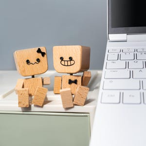 Op de afbeelding: Twee houten robotfiguren met lachende gezichten en vlinderdassen zitten in de buurt van een witte laptop. De robots zijn gemaakt van lichtgekleurd hout met zwarte details. Eén robot heeft een vlinderdas en de andere een strik op het hoofd. Het toetsenbord van de laptop is zichtbaar.
