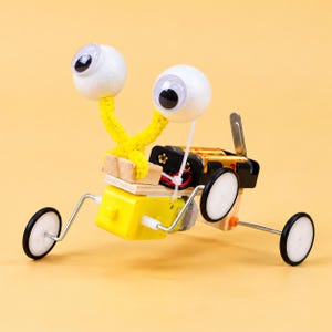 May include: A DIY motorized robot toy featuring two white foam ball eyes with black pupils on yellow pipe cleaner stalks. The body consists of a yellow motor, a black battery pack, wooden blocks, and four small white wheels on metal axles.