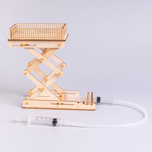 May include: A light-colored wooden scissor lift model featuring a top platform with a railing. The mechanism is operated by two clear plastic syringes connected by flexible tubing, allowing for hydraulic movement of the lift structure.