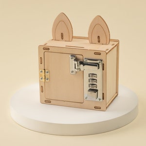 K&ouml;nnte beinhalten: Ein Sparschwein aus Holz in Form einer Box mit Zahlenschloss und zwei ohrenf&ouml;rmigen Aussparungen oben. Die Spardose hat eine Scharniert&uuml;r und ist aus hellem Holz gefertigt. Das Zahlenschloss ist silberfarben.