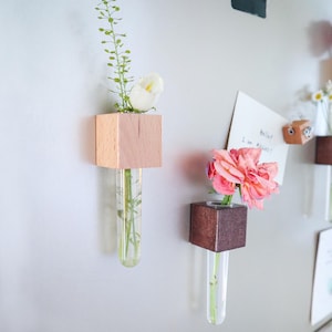 Könnte beinhalten: Holzwürfel-Vasen mit Glasröhren, die frische Blumen und Grünzeug enthalten. Eine Vase ist aus hellem Holz, die andere aus dunklem Holz. Die Vasen sind an einer Wand befestigt und bilden eine minimalistische Blumendekoration.