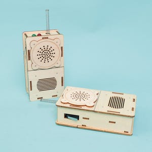 May include: Two wooden toy radios. One is upright with a spring antenna, red and green lights, and a speaker. The other is horizontal. Both have speaker holes and a light wood finish. The background is a light blue.