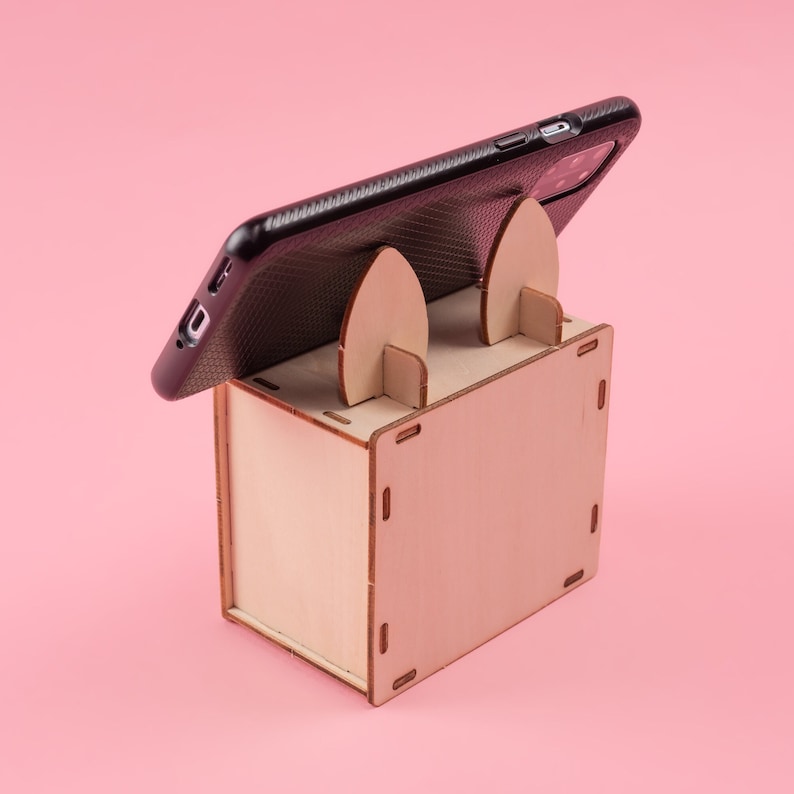 K&ouml;nnte beinhalten: Ein Handy-St&auml;nder aus hellem Holz mit einem schwarzen Telefon in einer H&uuml;lle. Der St&auml;nder hat zwei gebogene St&uuml;tzen. Das Telefon liegt auf dem St&auml;nder. Der Hintergrund ist einfarbig rosa.