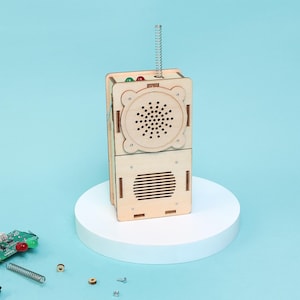 May include: A wooden toy radio with a spring antenna, red and green lights, and speaker holes. The radio is made of light-colored wood and sits on a white platform against a blue background. The radio has a speaker and a lower section with horizontal slots.
