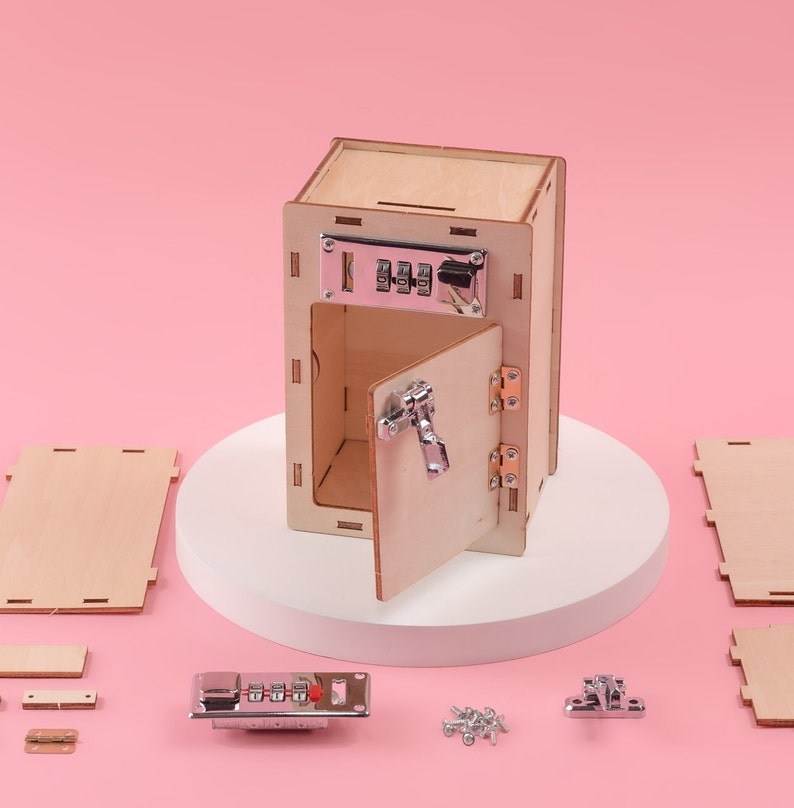 May include: A wooden toy safe with a combination lock, hinges, and a door that opens. The safe is on a white platform and there are various pieces of the safe scattered around it, including screws, hinges, and a combination lock.