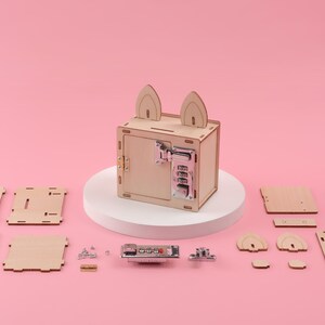 K&ouml;nnte beinhalten: Ein Holz-DIY-Tresor-Bausatz mit Zahlenschloss und Hasenohren-Design. Das Set enth&auml;lt verschiedene Holzteile, Schrauben und einen Zahlenschlossmechanismus. Der montierte Tresor steht auf einem wei&szlig;en Sockel vor einem rosa Hintergrund.
