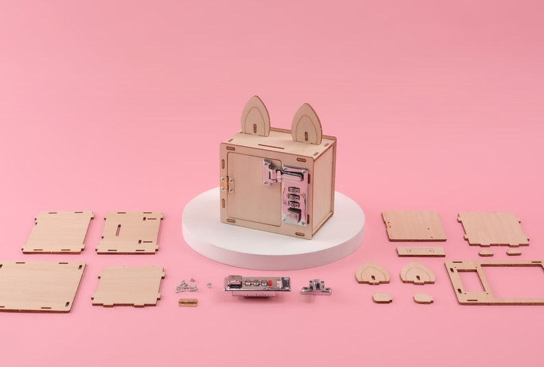 K&ouml;nnte beinhalten: Ein Holz-DIY-Tresor-Bausatz mit Zahlenschloss und Hasenohren-Design. Das Set enth&auml;lt verschiedene Holzteile, Schrauben und einen Zahlenschlossmechanismus. Der montierte Tresor steht auf einem wei&szlig;en Sockel vor einem rosa Hintergrund.