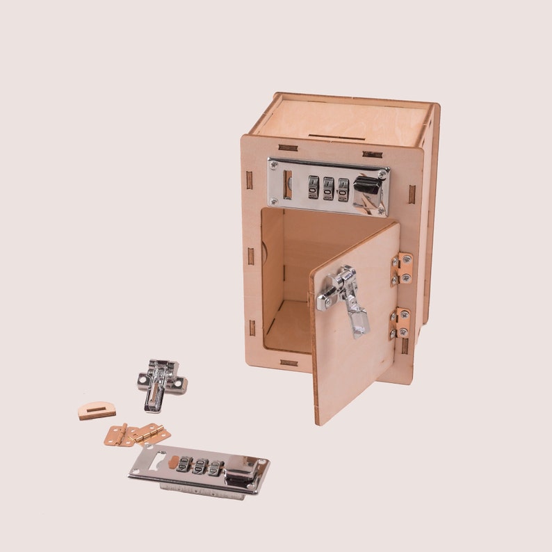 May include: A wooden toy safe with a combination lock, a latch, and a hinged door. The safe is open and the door is partially open. The safe is made of light wood and has a natural finish. The combination lock is silver and has three dials. The latch is silver and has a lever handle. The hinges are brass and are attached to the door and the safe. The safe is sitting on a white surface.