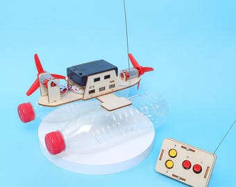 Peut inclure: Un kit de bateau télécommandé DIY composé d'un cadre en bois, deux bouteilles en plastique transparent avec bouchons rouges, deux hélices rouges, un boîtier à piles noir et une télécommande en bois rectangulaire avec deux boutons jaunes et deux rouges.